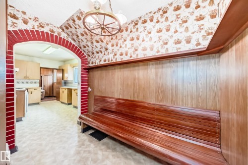 Built-in wooden bench with slatted details, wood-paneled walls, decorative wallpaper, brick archway, and overhead wagon wheel light fixture - 11929 91 Street, Edmonton, AB 