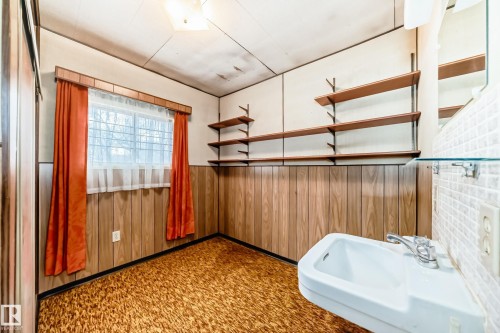 Room featuring wood-paneled wainscoting, wall-mounted shelving, textured flooring, and a window with privacy sheers - 11929 91 Street, Edmonton, AB 