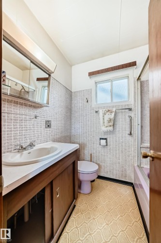 Bathroom featuring a wood-finish vanity with an integrated sink, textured wall tiling, patterned flooring, and a window with frosted glass - 11929 91 Street, Edmonton, AB 