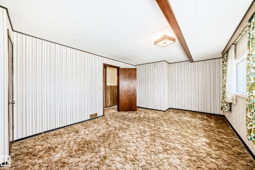Interior room featuring patterned carpeting, striped wall paneling, a ceiling beam, a flush-mount light fixture, and a window with floral drapes - 11929 91 Street, Edmonton, AB 