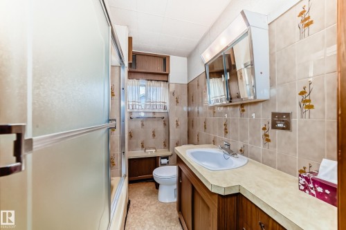 Full bathroom featuring a frosted glass tub/shower enclosure, wood-finish vanity with an integrated sink and light-colored countertop, wall-mounted mirrored medicine cabinet, and tiled walls with decorative accents - 11929 91 Street, Edmonton, AB 