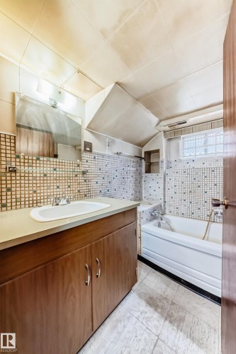 Bathroom featuring a wood-finish vanity with a single basin sink, a tiled backsplash, a bathtub with a shower fixture, and patterned floor tiling - 11929 91 Street, Edmonton, AB 