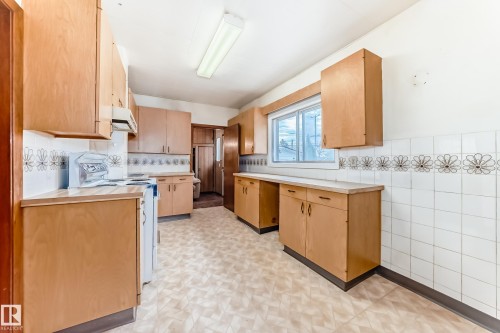 Spacious kitchen featuring extensive wood-finish cabinetry, a large window, tiled backsplash with decorative accents, an overhead fluorescent light fixture, and patterned sheet flooring - 11929 91 Street, Edmonton, AB 