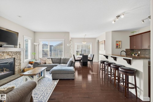Open-concept living area featuring rich wood-finish flooring, a stone-clad fireplace with a hearth, multiple windows with blinds, a spacious breakfast bar with built-in supports, and recessed lighting - 8003 Summerside Grande Boulevard, Edmonton, AB - Indoor Photo Showing Living Room With Fireplace