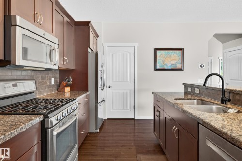 Kitchen featuring rich wood-finish cabinetry, stainless steel appliances, speckled stone countertops, and a tile backsplash - 8003 Summerside Grande Boulevard, Edmonton, AB - Indoor Photo Showing Kitchen With Double Sink