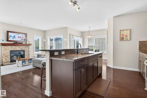 Kitchen island featuring dark wood-finish cabinetry, a stone countertop, and an undermount sink with a matte black faucet - 8003 Summerside Grande Boulevard, Edmonton, AB - Indoor Photo Showing Kitchen With Fireplace