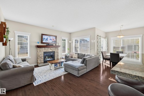 Spacious open-concept living area featuring dark wood-finish flooring, a stone-faced fireplace with a wood mantel, and multiple windows with blinds - 8003 Summerside Grande Boulevard, Edmonton, AB - Indoor Photo Showing Living Room With Fireplace