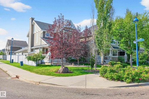 Corner property featuring a light-toned exterior with stone accents, an inviting front porch with white railings, and mature trees providing natural shading - 8003 Summerside Grande Boulevard, Edmonton, AB - Outdoor