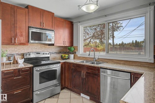 Kitchen featuring wood cabinetry, stainless steel appliances, a double basin sink with a gooseneck faucet, light-toned countertops, and a decorative tile backsplash - 16715 79A Avenue, Edmonton, AB - Indoor Photo Showing Kitchen With Double Sink