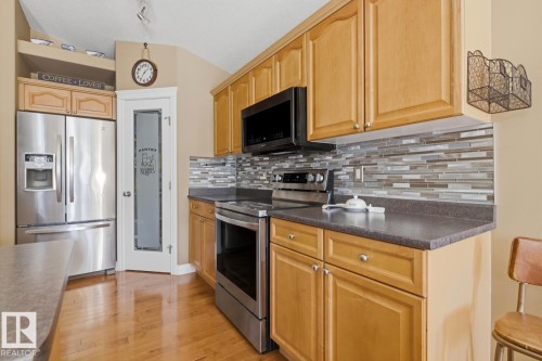 Kitchen featuring stainless steel appliances, wood-finish flooring, a mosaic tile backsplash, light wood cabinetry, and a pantry with a frosted glass door - 9 Danfield Place, Spruce Grove, AB - Indoor Photo Showing Kitchen