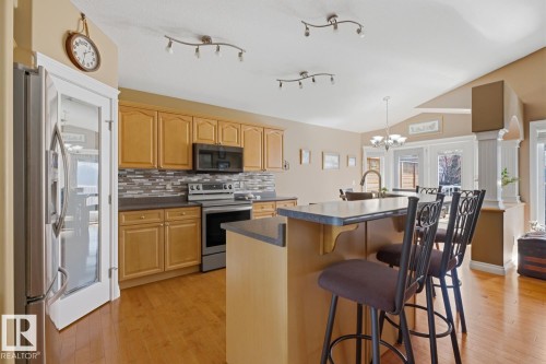 Open-concept kitchen featuring wood-finish flooring, light wood cabinetry, stainless steel appliances, a tiled backsplash, and an island with bar seating - 9 Danfield Place, Spruce Grove, AB - Indoor Photo Showing Kitchen