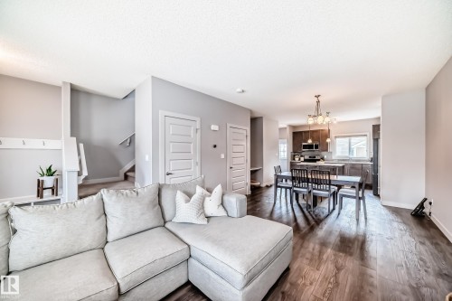Open-concept living area featuring wood-finish flooring, a contemporary kitchen with dark wood cabinetry, and a dining area illuminated by a modern chandelier - 6919 Cardinal Wynd, Edmonton, AB - Indoor Photo Showing Living Room