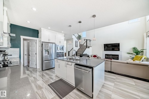 Open-concept living space featuring a central kitchen island with stainless steel appliances, light wood-finish flooring, a contemporary fireplace, and a striking dark accent wall - 3716 46 Avenue, Beaumont, AB - Indoor Photo Showing Kitchen With Double Sink With Upgraded Kitchen