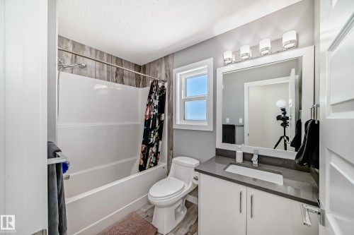 Full bathroom featuring a white tub and shower combination with a textured grey tile surround, a single-pane window, and wood-finish flooring - 3716 46 Avenue, Beaumont, AB - Indoor Photo Showing Bathroom