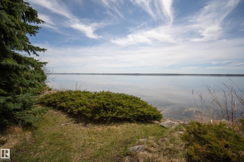 Expansive waterfront featuring a calm body of water, a natural shoreline, mature evergreen trees, and native groundcover - 77 Lakeshore Drive, Rural Leduc County, AB - Outdoor With Body Of Water With View