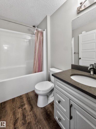 The bathroom features a white tub and shower combination, a white toilet, and a vanity with a dark countertop and white cabinetry - 15 2072 Wonnacott Way, Edmonton, AB - Indoor Photo Showing Bathroom