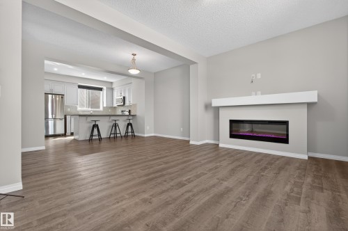 Spacious living area featuring durable flooring, an electric fireplace with a white mantel, and an open layout to the kitchen with a breakfast bar - 15 2072 Wonnacott Way, Edmonton, AB - Indoor Photo Showing Living Room With Fireplace
