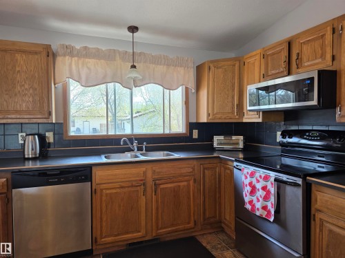 Kitchen featuring wood cabinetry, dark countertops, dark tile backsplash, a double basin sink, and stainless steel appliances including a dishwasher, range, and microwave - 4029 55 Avenue, Drayton Valley, AB - Indoor Photo Showing Kitchen With Double Sink