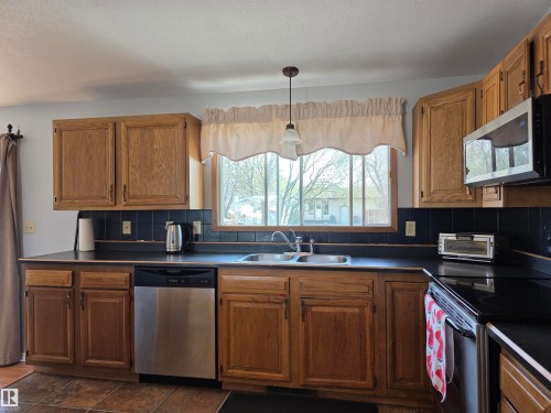 Kitchen featuring wood cabinetry, black countertops, a tile backsplash, stainless steel appliances, and a double basin sink beneath a window - 4029 55 Avenue, Drayton Valley, AB - Indoor Photo Showing Kitchen With Double Sink