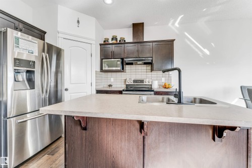 Kitchen featuring an island with an integrated sink and black faucet, stainless steel appliances, dark wood-finish cabinetry, and a subway tile backsplash - 2292 Casey Crescent, Edmonton, AB - Indoor Photo Showing Kitchen With Upgraded Kitchen