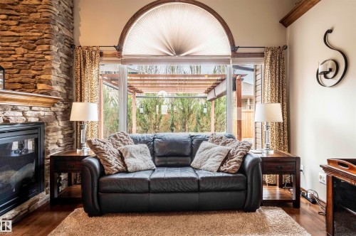 Living area featuring a stone-clad fireplace with a wood mantel, wood-finish flooring, a full-height window with an arched transom, and an outdoor pergola structure - 19 Newton Place, St. Albert, AB - Indoor Photo Showing Living Room With Fireplace