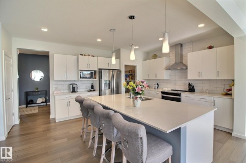 Modern kitchen featuring a large center island with seating, white shaker cabinetry, stainless steel appliances, a subway tile backsplash, and wood-finish flooring - 121 Stiles Bend, Leduc, AB - Indoor Photo Showing Kitchen With Upgraded Kitchen