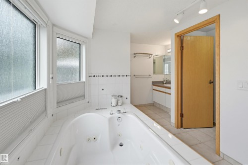 White tiled bathroom featuring a built-in jetted tub, two large privacy windows with blinds, track lighting, and a separate vanity area with a dual-sink setup and white cabinetry - 317 Hollick-Kenyon Road, Edmonton, AB - Indoor