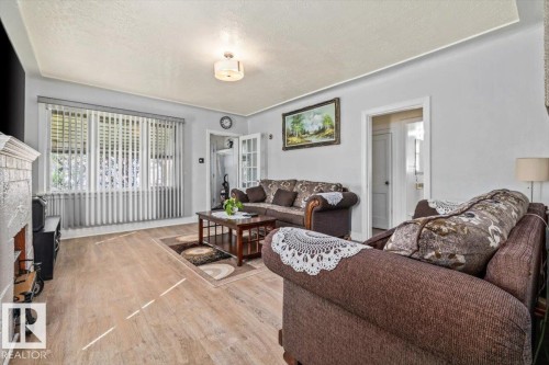 Living area featuring light wood-finish flooring, a decorative fireplace with a stone-finish facade, a large window with vertical blinds, and a neutral color palette - 10939 96 Street, Edmonton, AB - Indoor Photo Showing Living Room