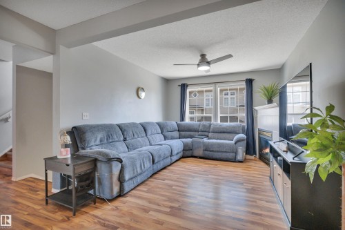 Living area featuring wood-finish flooring, a ceiling fan, a fireplace with a white mantel, and windows with blue drapery - 102 4415 South Park Drive, Leduc, AB - Indoor Photo Showing Living Room