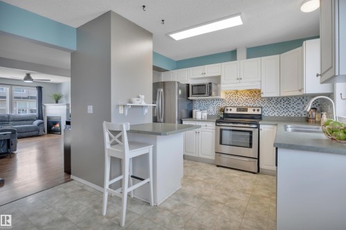 Kitchen featuring white cabinetry, stainless steel appliances, a mosaic tile backsplash, and a breakfast bar with a solid surface countertop - 102 4415 South Park Drive, Leduc, AB - Indoor Photo Showing Kitchen With Double Sink With Upgraded Kitchen