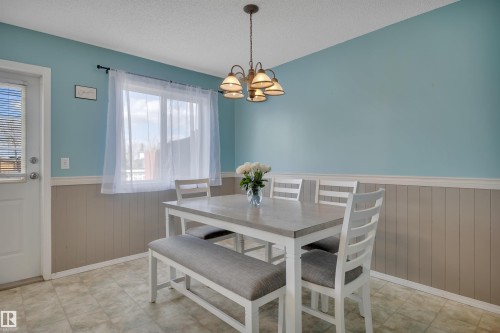 Dining area featuring a ceiling-mounted chandelier, window with privacy blinds, blue painted walls, beadboard wainscoting, and patterned vinyl flooring - 102 4415 South Park Drive, Leduc, AB - Indoor Photo Showing Dining Room