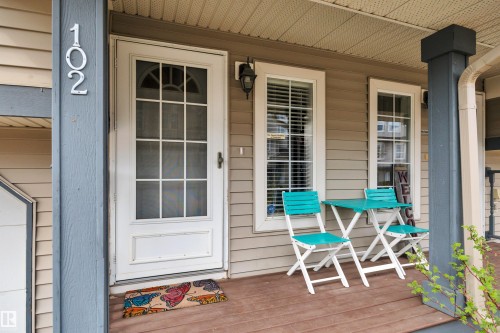 Inviting front porch featuring wood-finish flooring, a white glass-paned storm door, and two framed windows with horizontal blinds - 102 4415 South Park Drive, Leduc, AB - Outdoor With Deck Patio Veranda With Exterior