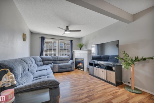 Inviting living space featuring wood-finish flooring, a white-mantled fireplace, and a ceiling fan with integrated lighting - 102 4415 South Park Drive, Leduc, AB - Indoor Photo Showing Living Room With Fireplace