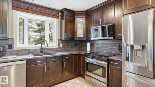 Kitchen featuring rich wood cabinetry, stainless steel appliances, a bay window, dark countertop, and a tiled backsplash - 27 Addison Crescent, St. Albert, AB - Indoor Photo Showing Kitchen With Double Sink With Upgraded Kitchen