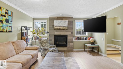 Living area featuring a modern fireplace with a shiplap surround, wood-finish flooring, two large windows, a built-in bookshelf, and crown molding - 27 Addison Crescent, St. Albert, AB - Indoor Photo Showing Living Room With Fireplace