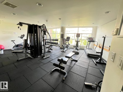 Fitness area featuring resilient rubber flooring, a multi-station weight machine, cardio equipment, and natural light from large windows - 710 2504 109 Street Nw, Edmonton, AB 