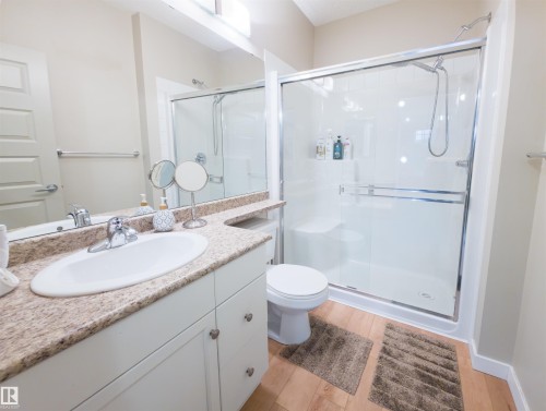 Bathroom featuring a single vanity with an integrated sink and granite-finish countertop, white base cabinetry with brushed nickel hardware, a mirrored wall, and a glass-enclosed shower with white surround and built-in bench - 135 7825 71 Street, Edmonton, AB - Indoor Photo Showing Bathroom