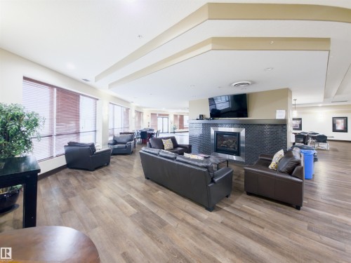Spacious common area featuring wood-finish flooring, multiple large windows, and a central fireplace with dark tile surround - 135 7825 71 Street, Edmonton, AB - Indoor With Fireplace
