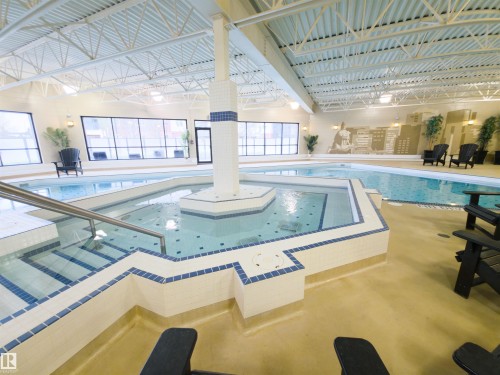 Indoor aquatic center featuring a multi-level pool with integrated spa, mosaic tile detailing, large windows, and an exposed truss ceiling - 135 7825 71 Street, Edmonton, AB - Indoor Photo Showing Other Room With In Ground Pool