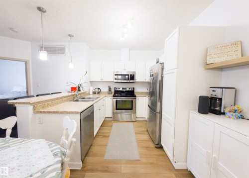 Bright kitchen featuring white cabinetry, stainless steel appliances, light-toned countertops, wood-finish flooring, and pendant lighting over a breakfast bar - 135 7825 71 Street, Edmonton, AB - Indoor Photo Showing Kitchen With Stainless Steel Kitchen With Double Sink
