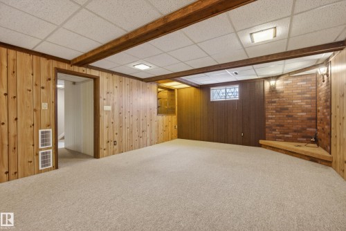 Spacious room featuring light-toned carpeting, wood paneling, exposed ceiling beams, a brick accent wall with sconce lighting, and a built-in corner platform - 12016 51 Street, Edmonton, AB - Indoor Photo Showing Basement
