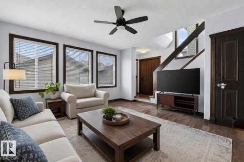 Living area featuring wood-finish flooring, a contemporary ceiling fan, and three large windows with dark trim - 108 Hewitt Circle, Spruce Grove, AB - Indoor Photo Showing Living Room