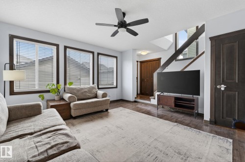 Living area featuring dark wood-finish flooring, three large windows with blinds, a dark ceiling fan, a built-in staircase, and a dark wood door - 108 Hewitt Circle, Spruce Grove, AB - Indoor Photo Showing Living Room