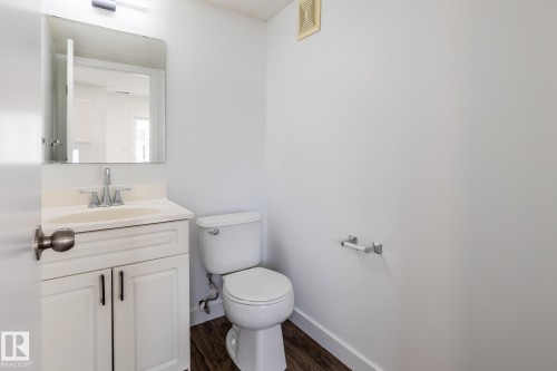 Bathroom featuring a white vanity with integrated sink, chrome faucet, wall-mounted mirror with overhead lighting, white toilet, and wood-finish flooring - #136, 180 Bridgeport Bv, Leduc, AB - Indoor Photo Showing Bathroom