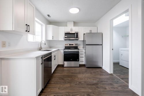 Contemporary kitchen featuring white cabinetry, light countertops, wood-finish flooring, and stainless steel appliances including a refrigerator, range, microwave, and dishwasher - #136, 180 Bridgeport Bv, Leduc, AB - Indoor Photo Showing Kitchen