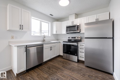 Modern kitchen featuring white shaker cabinetry, stainless steel appliances, and a wood-finish flooring - #136, 180 Bridgeport Bv, Leduc, AB - Indoor Photo Showing Kitchen