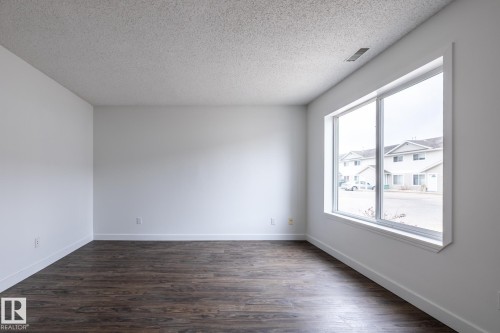 Spacious room featuring white walls, deep brown wood-finish flooring, white baseboards, and a large multi-pane window - #136, 180 Bridgeport Bv, Leduc, AB - Indoor Photo Showing Other Room