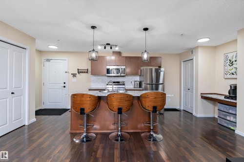 Open-concept kitchen featuring wood-finish flooring, a central island with a granite-style countertop, stainless steel appliances, and pendant lighting - 122 3357 16A Avenue, Edmonton, AB - Indoor Photo Showing Kitchen With Upgraded Kitchen