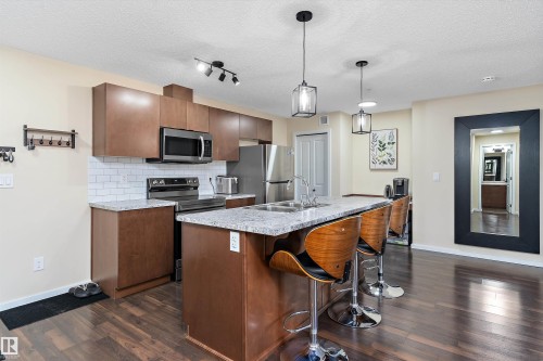 Open-concept kitchen featuring wood-finish flooring, stainless steel appliances, a white subway tile backsplash, light-toned countertops, and a kitchen island with pendant lighting - 122 3357 16A Avenue, Edmonton, AB - Indoor Photo Showing Kitchen With Double Sink With Upgraded Kitchen