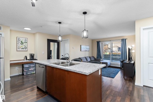 Open-concept living space featuring wood-finish flooring, a kitchen island with dual sinks and a stainless steel dishwasher, and a built-in countertop desk - 122 3357 16A Avenue, Edmonton, AB - Indoor Photo Showing Kitchen With Double Sink